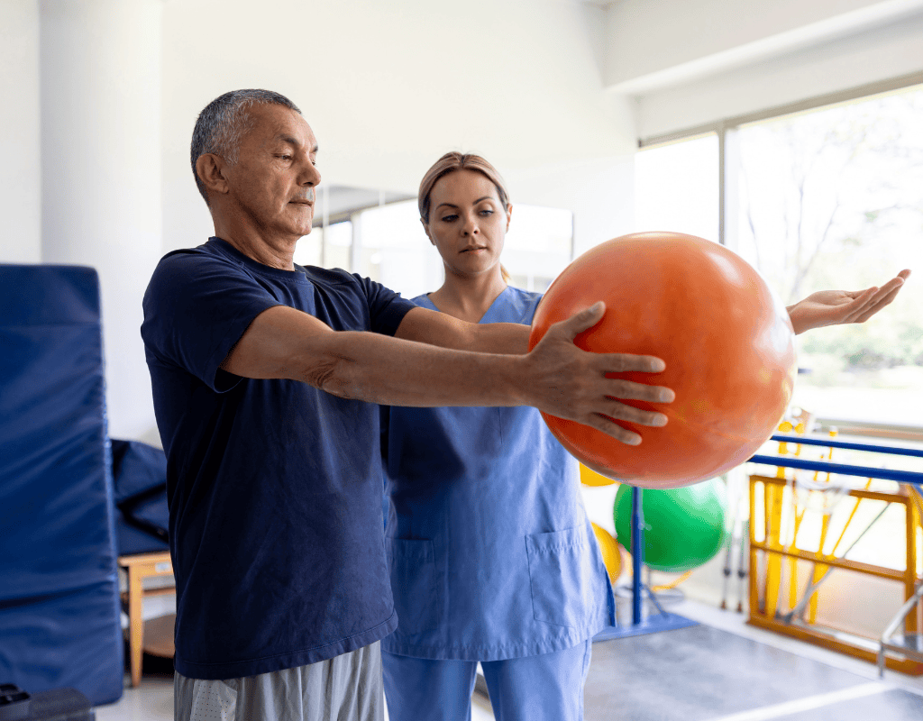 Therapist and an older man holding a large orange exercise ball together in a clinic gym