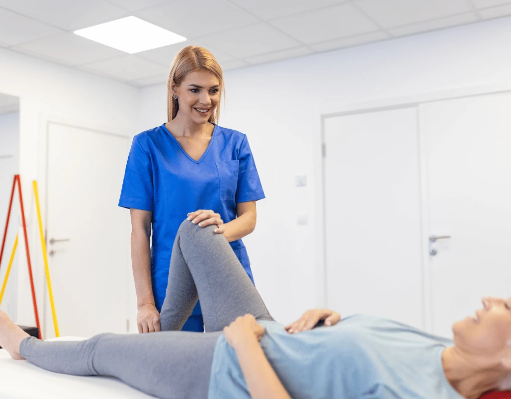Therapist in blue scrubs raising and stretching a patients leg while she lies on a treatment table