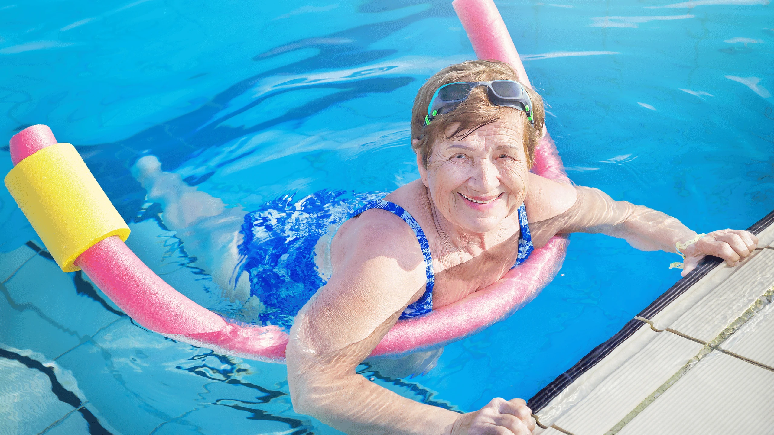 Older woman in goggles smiling while holding a pink pool noodle in a swimming pool