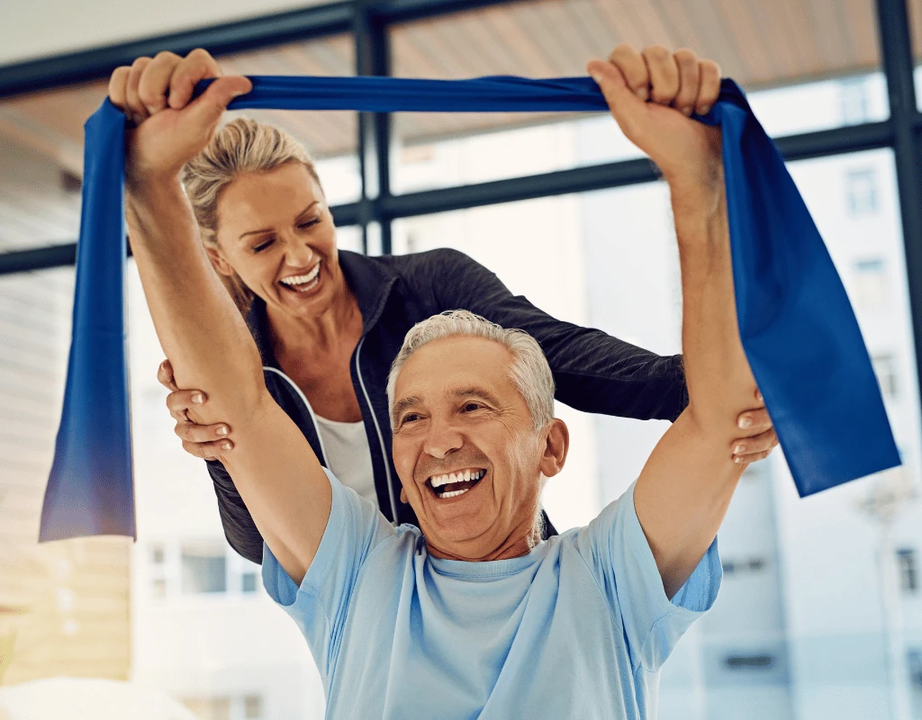 Therapist helping a smiling older man raise a blue resistance band overhead with both hands