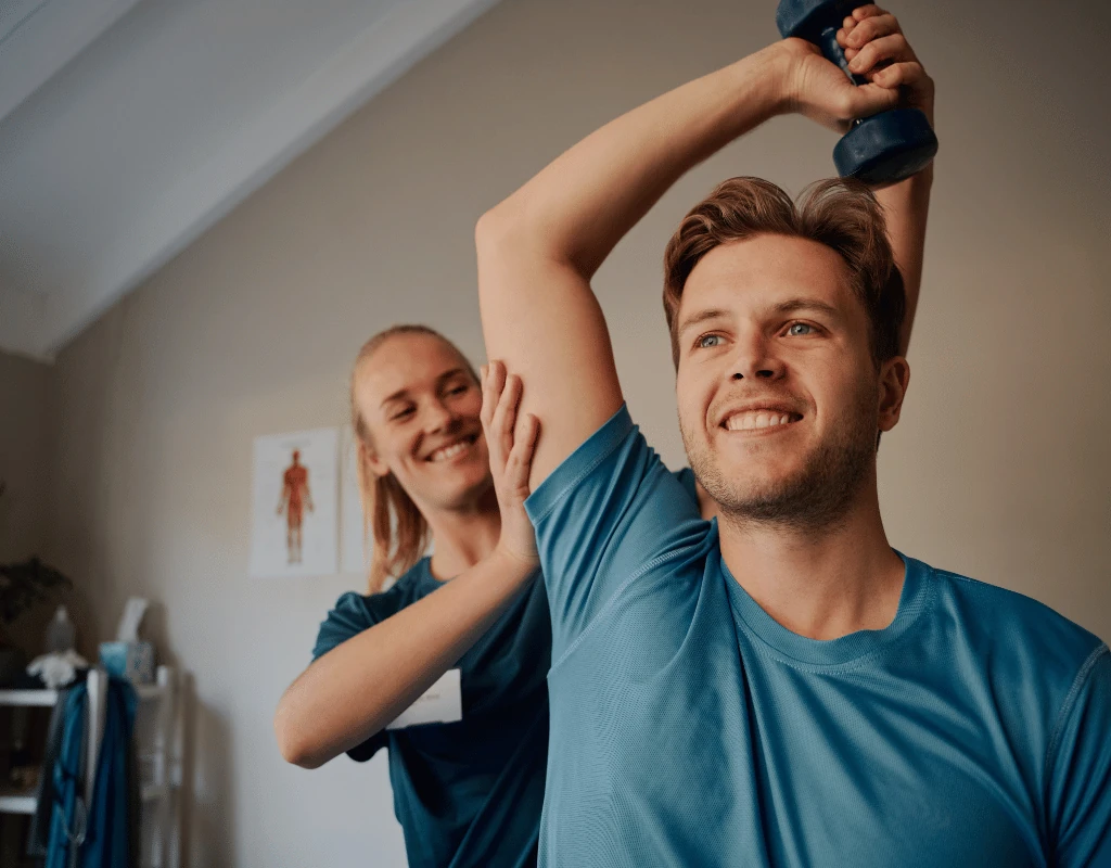 Therapist standing behind a young man supporting his arms through an overhead dumbbell press