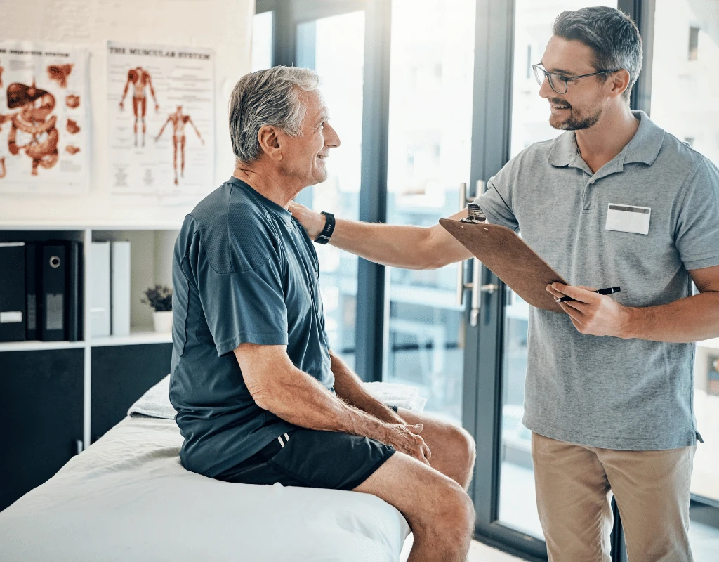 Clinician holding a clipboard talking with an older man seated on a treatment table