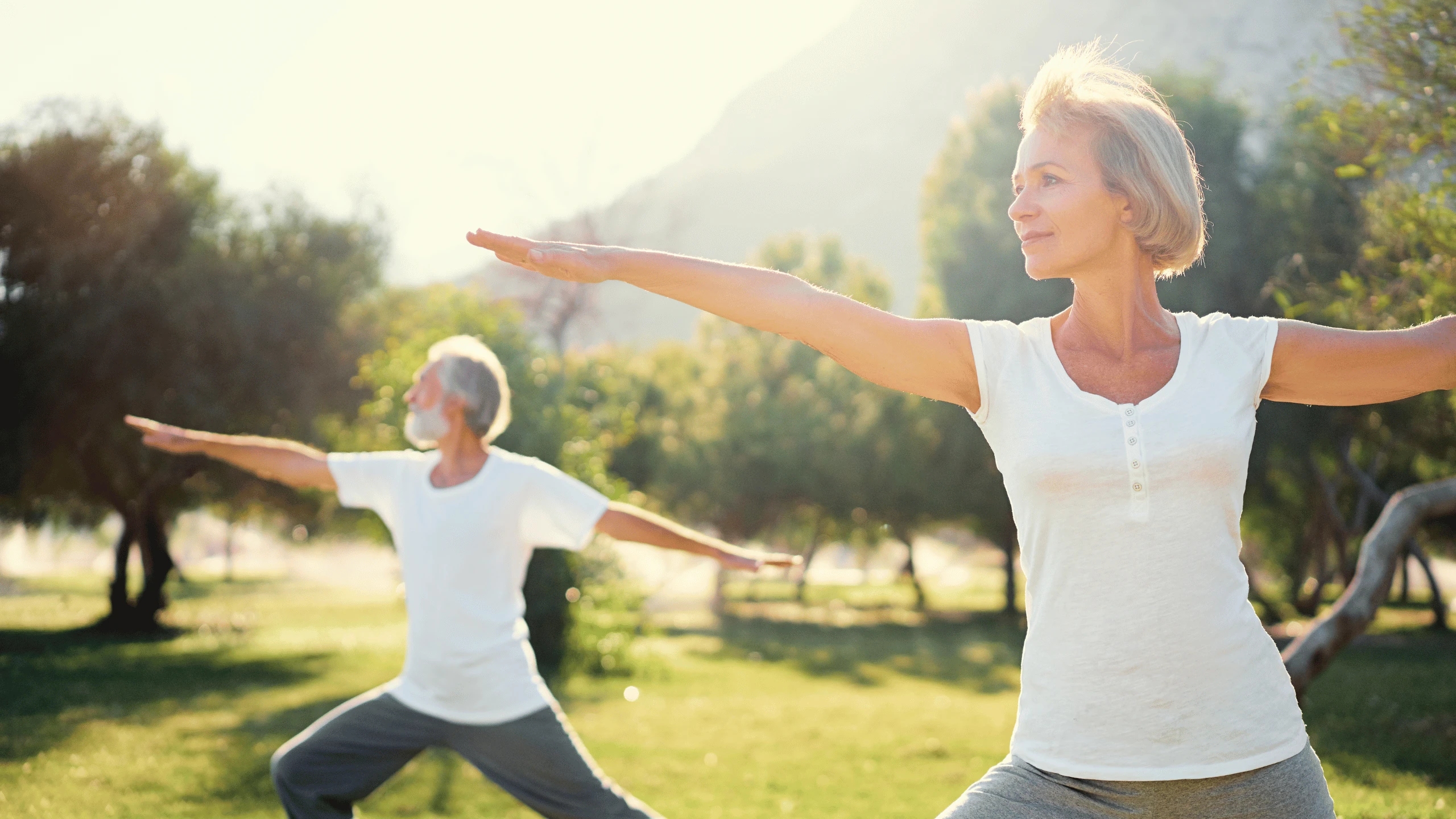 Two older women standing in a wide-legged yoga pose with arms outstretched on grass