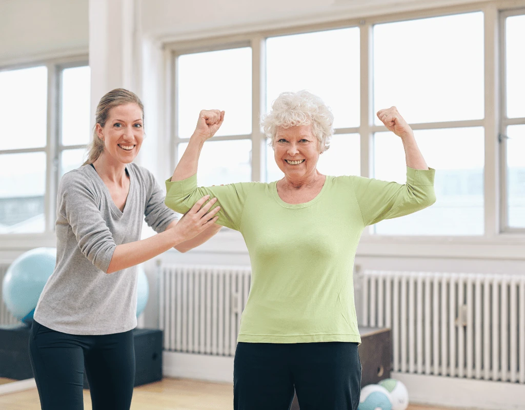 Therapist and a smiling older woman both flexing their arms in a bright therapy gym