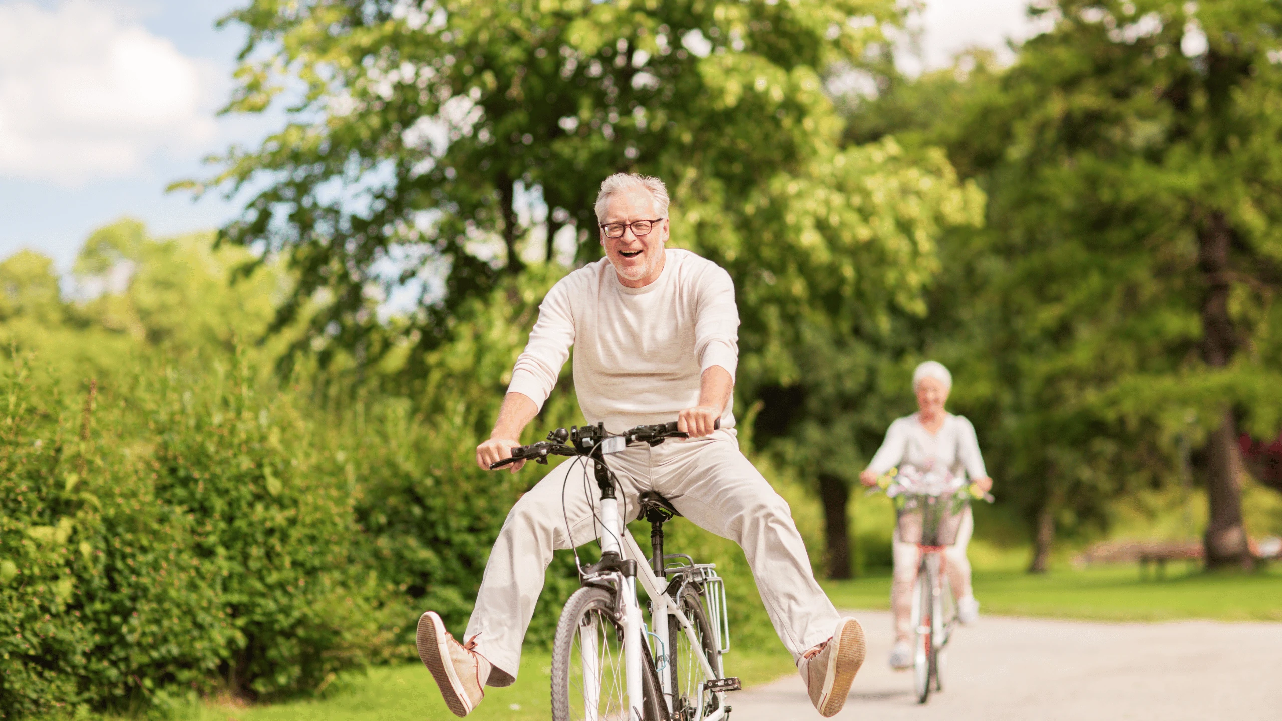 Older man and woman riding bicycles along a paved park path lined with trees