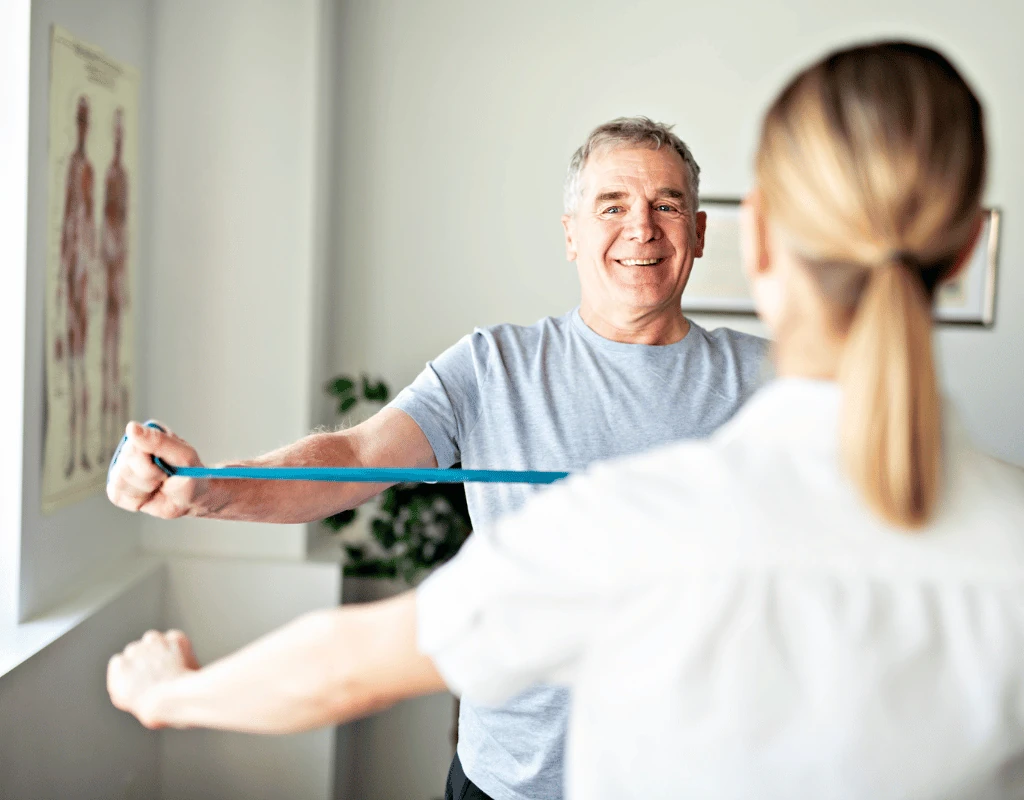 Man pulling a blue resistance band towards his chest while a therapist stands facing him