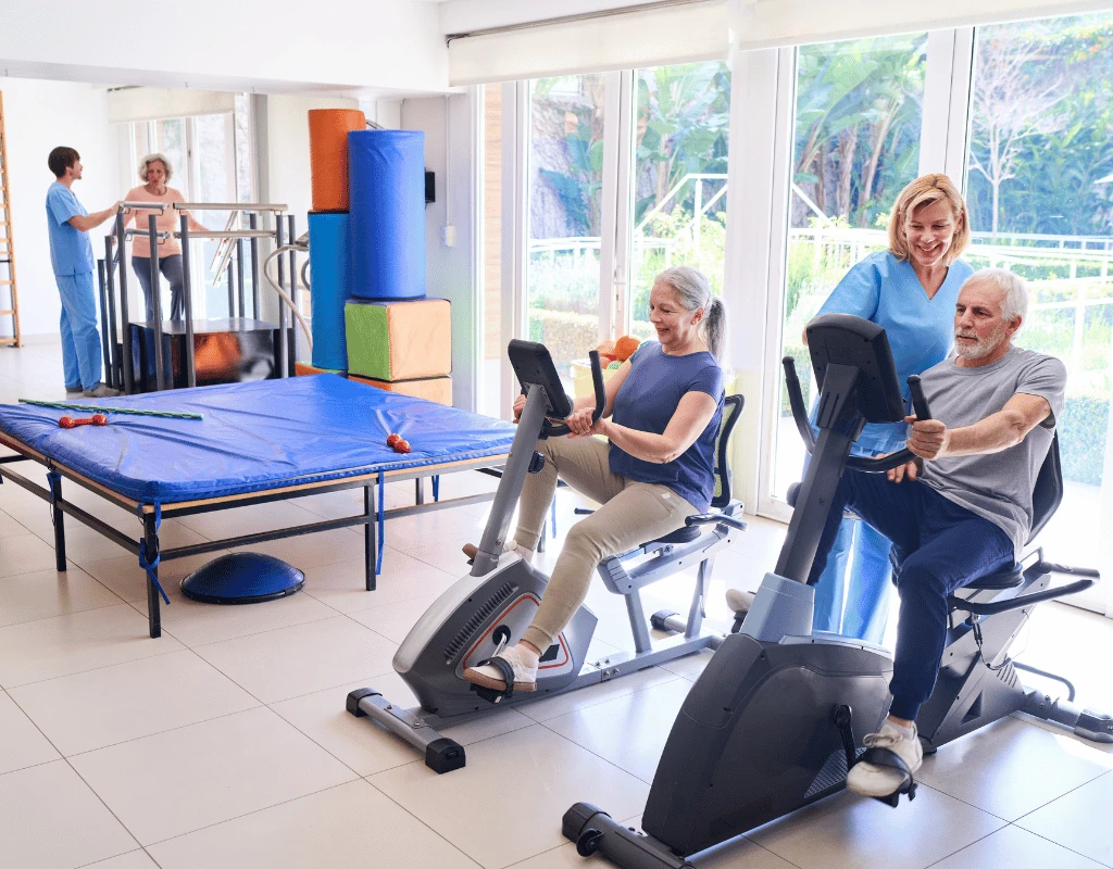 Open therapy gym containing exercise bikes, parallel bars and treatment tables with patients working out