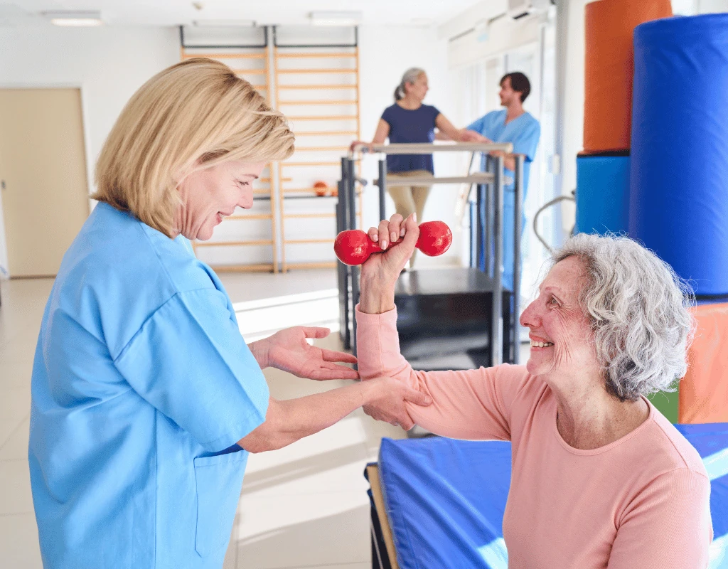 Therapist kneeling beside a seated older woman guiding her arm through a dumbbell exercise