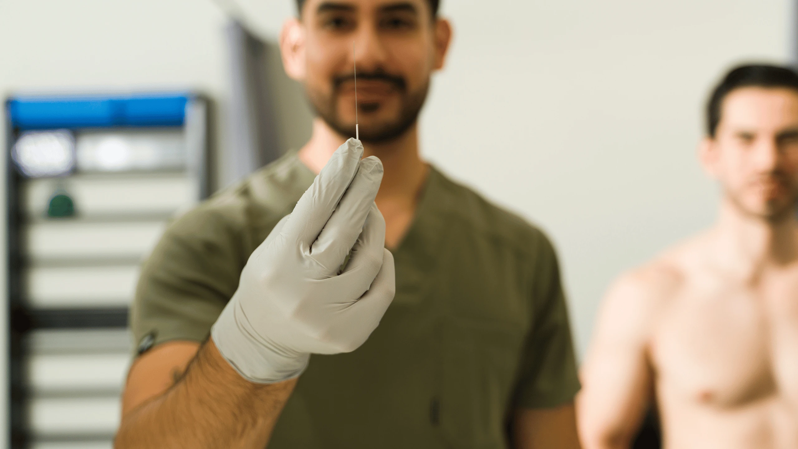 Gloved clinician holding a fine needle with a shirtless male patient seated in the background