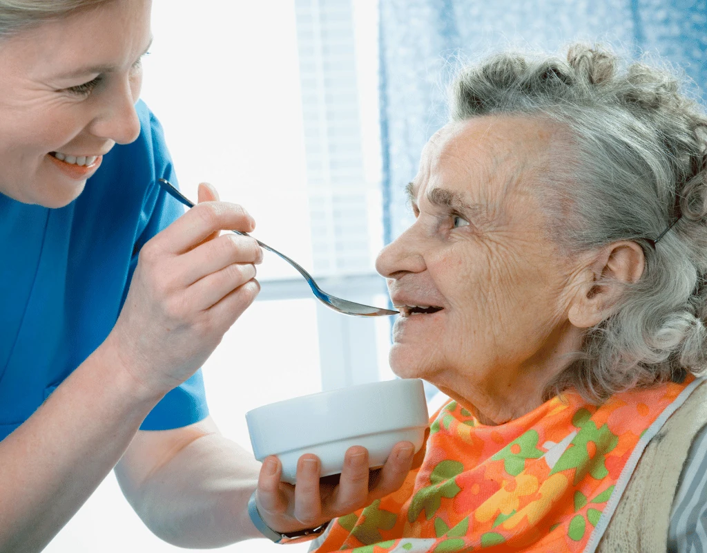 Clinician in blue scrubs holding a bowl and offering a spoonful of food to an elderly woman
