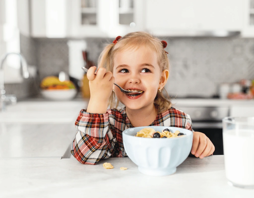 Young girl in a checked shirt eating cereal from a bowl at a kitchen counter