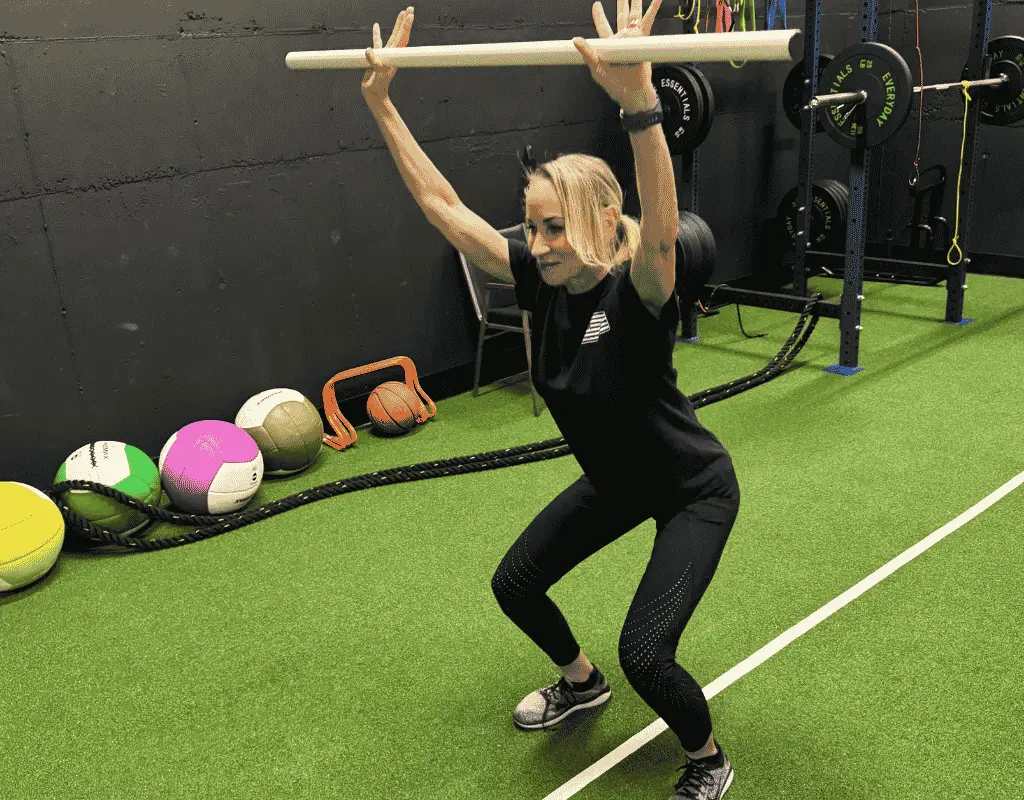 Woman holding a wooden dowel overhead with both arms extended while squatting deeply on turf