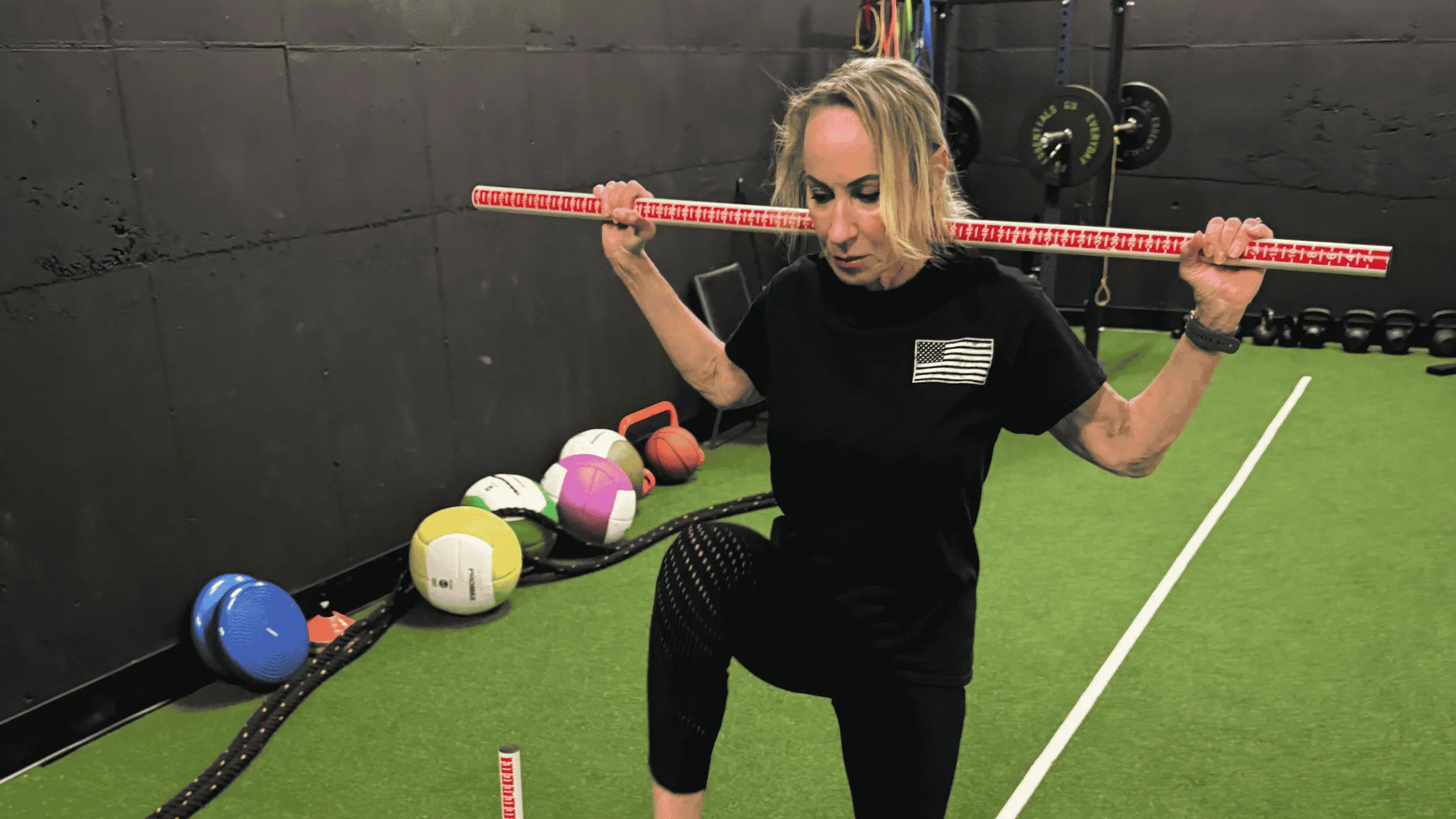 Woman in black activewear holding a wooden dowel across her shoulders while lowering into a squat on indoor turf