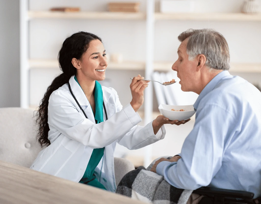 Clinician in a white coat offering a spoonful of food to an older man seated in a chair