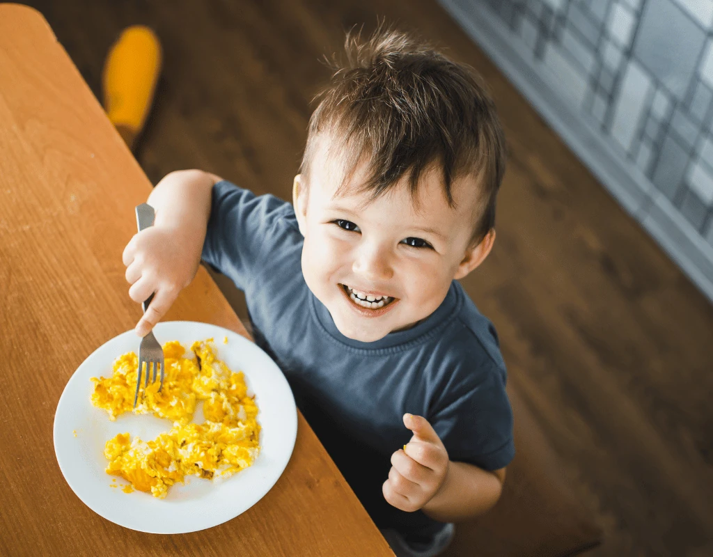 Smiling toddler seated at a table with a fork beside a plate of scrambled eggs