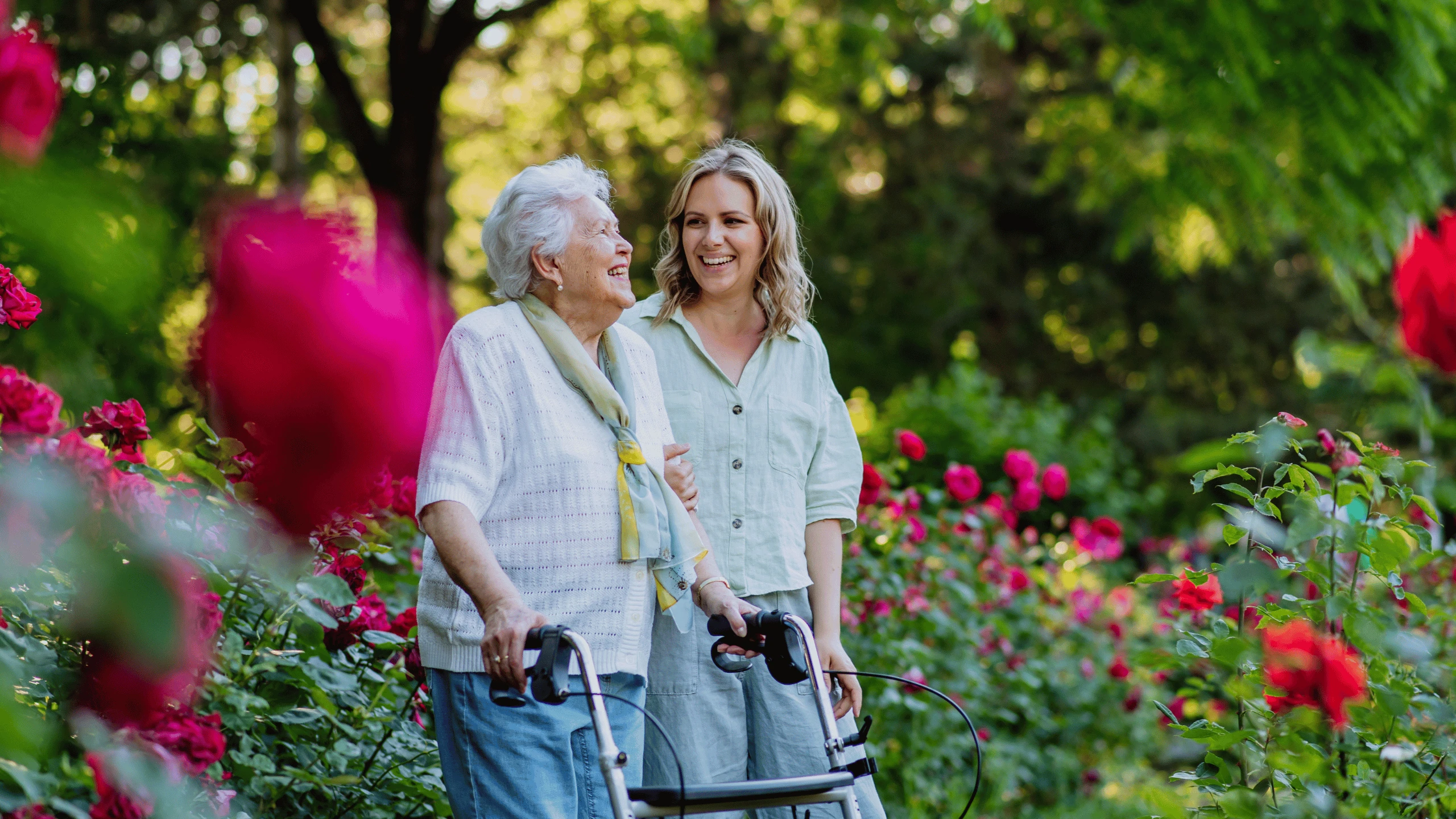 Two women walking along a flower-lined garden path, one using a rolling walker