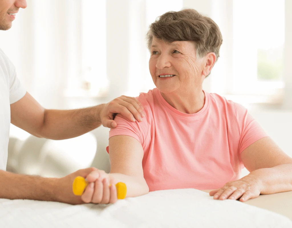 Therapists hand on an older womans shoulder as she lifts a yellow dumbbell resting on a table