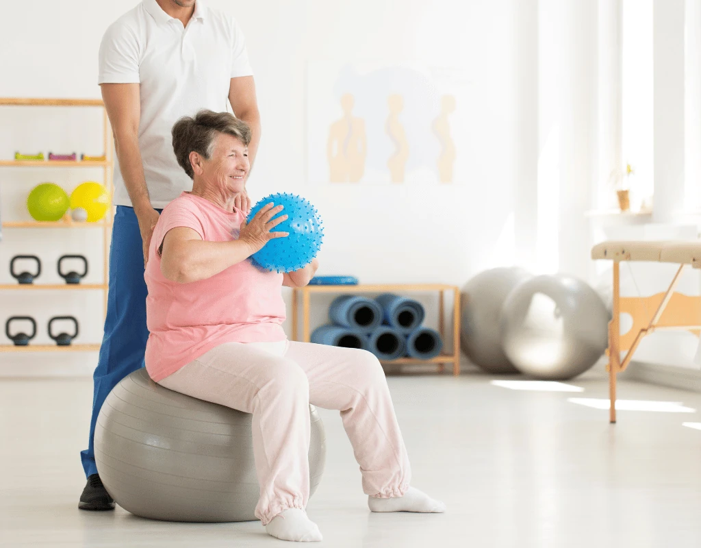 Therapist guiding an older woman holding a blue ball while she sits on a grey exercise ball