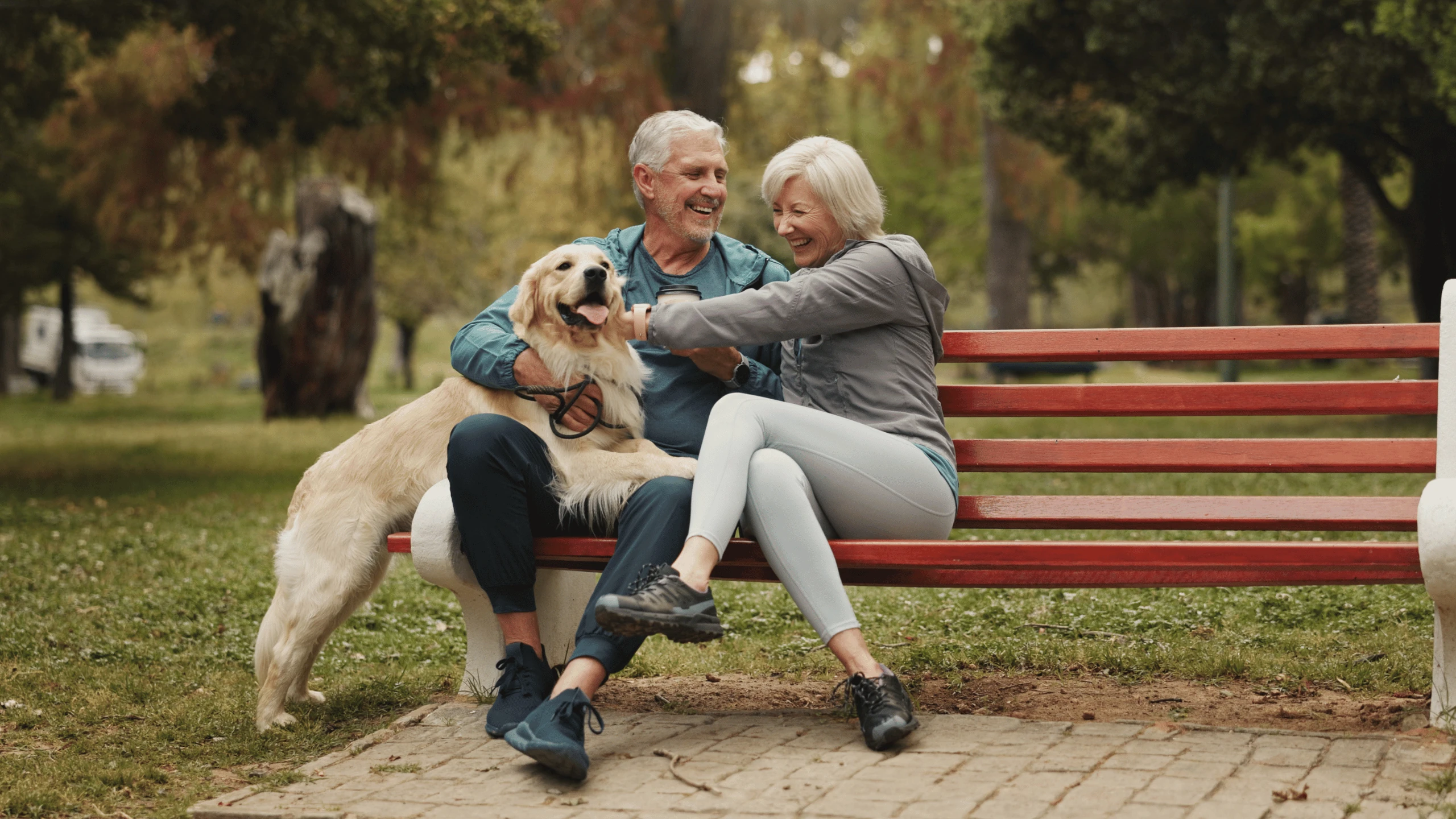 Older man and woman sitting on a park bench reaching down to pet a dog