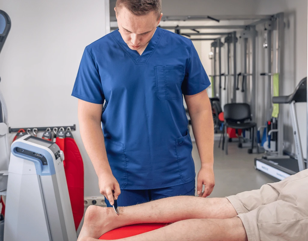 Therapist in blue scrubs treating a patients leg on a red treatment table with gym equipment behind