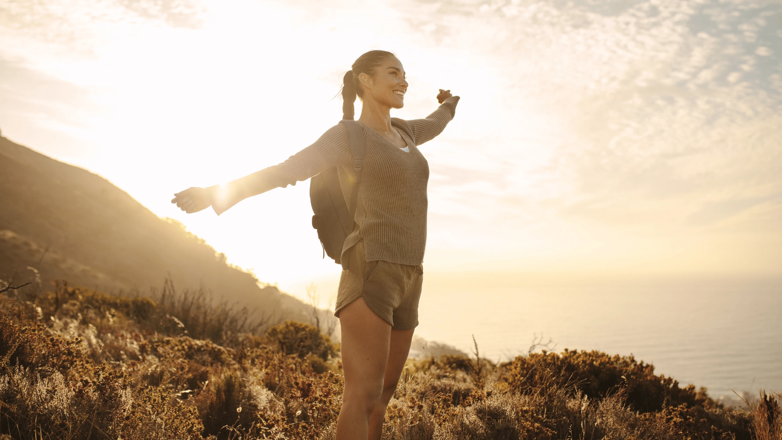 Woman standing on a hilltop with arms outstretched against a sunset sky