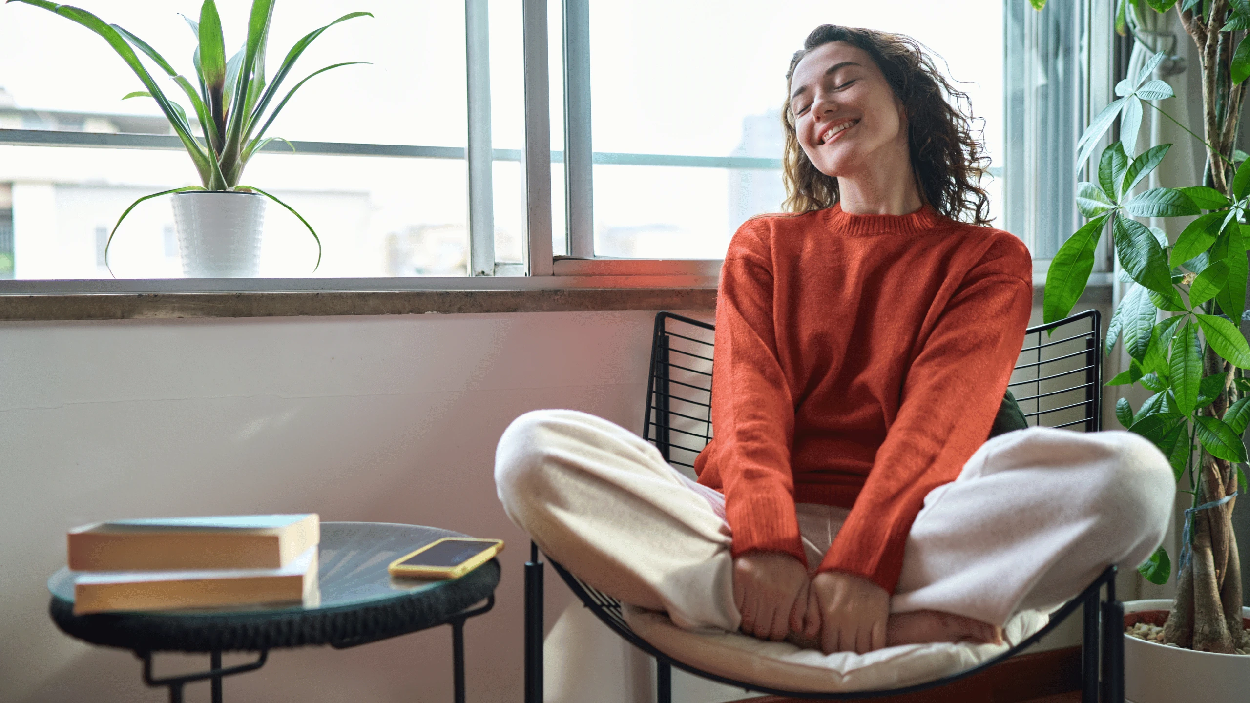 Woman sitting cross-legged in a chair by a window with her arms relaxed and eyes closed