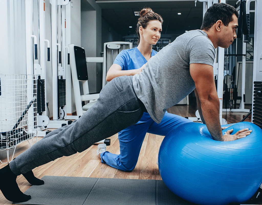 Therapist supporting a man holding a plank position with his hands on a blue stability ball in a gym