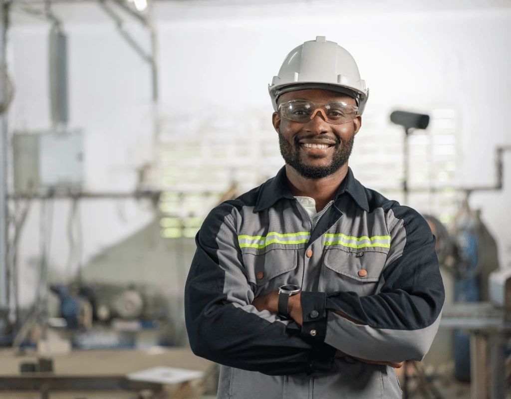 Worker in a white hard hat and safety glasses standing with arms folded in an industrial workshop