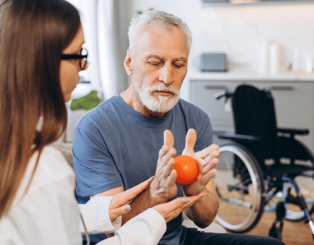 Therapist guiding an older man squeezing an orange therapy ball, a wheelchair visible behind them