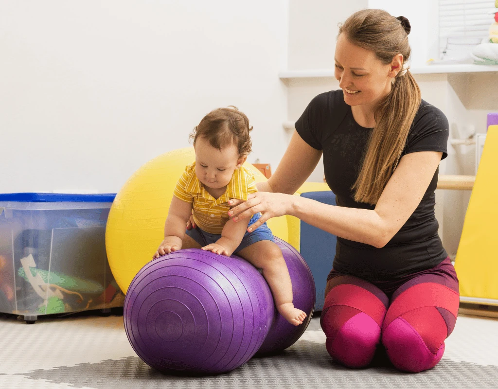 Therapist supporting a baby balanced on a purple therapy ball while the mother kneels alongside