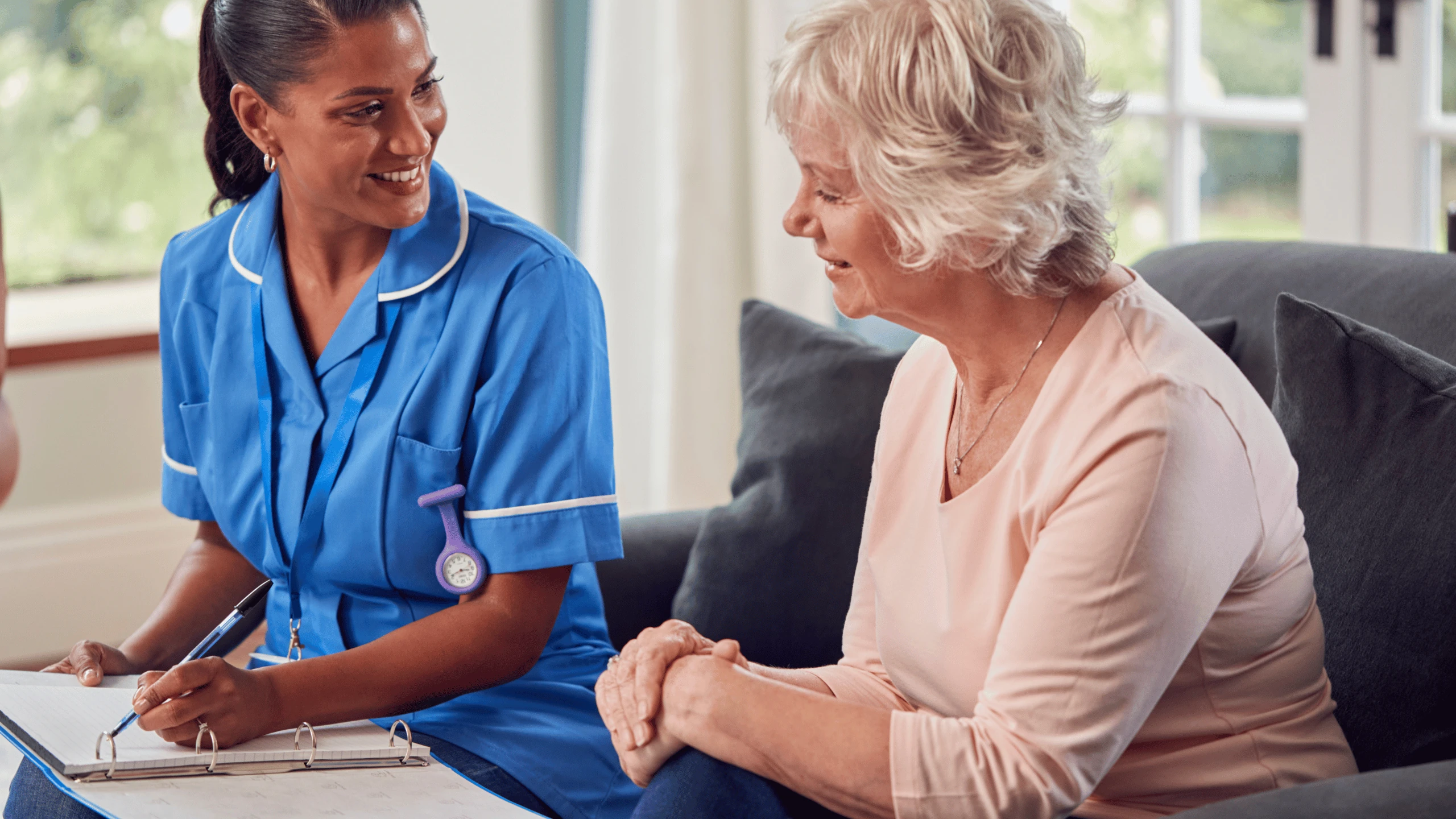 Clinician with a clipboard sitting beside an older woman in a living room