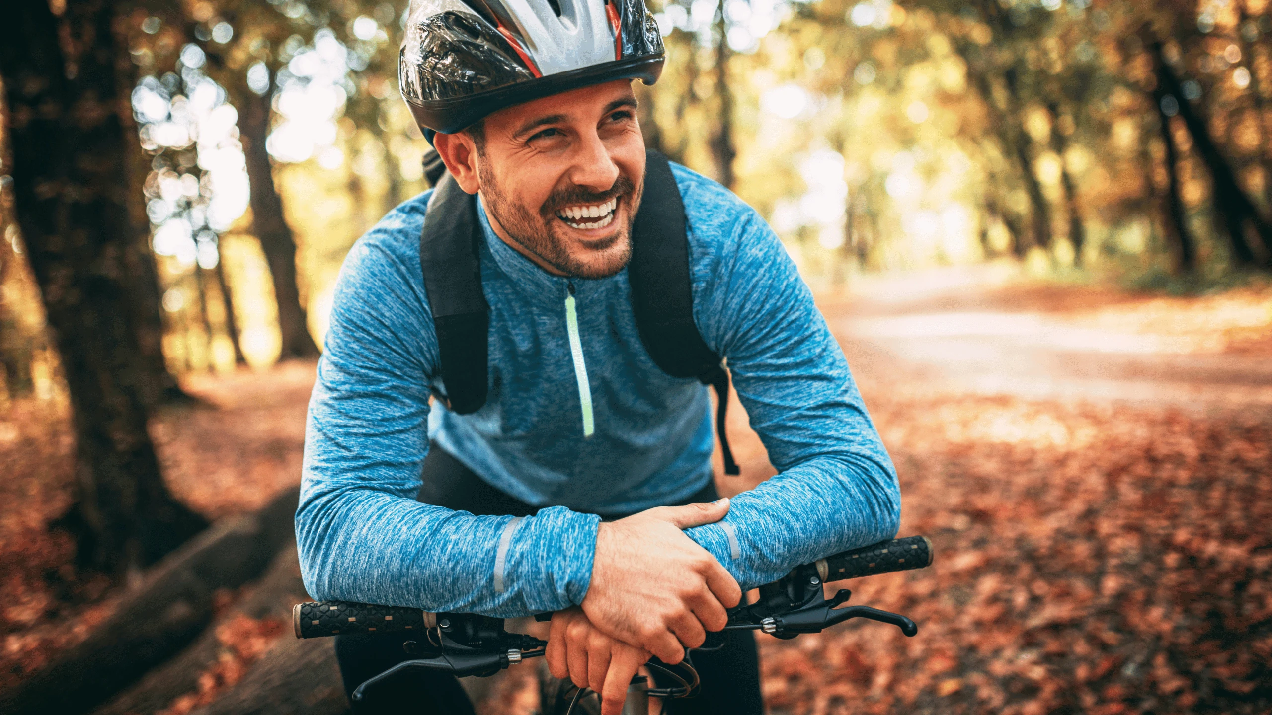 Man in a cycling helmet and jacket leaning on his handlebars on a leaf-covered trail