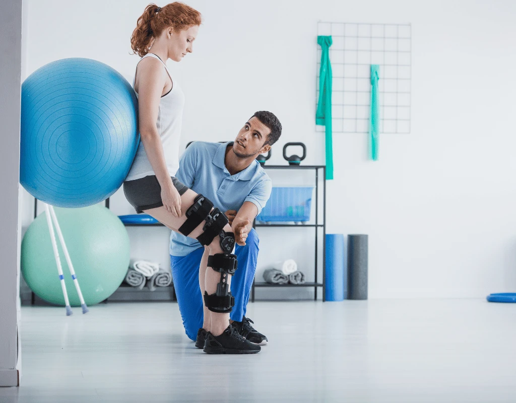 Man kneeling on a gym floor adjusting a knee brace on his leg while a woman with an exercise ball works behind him