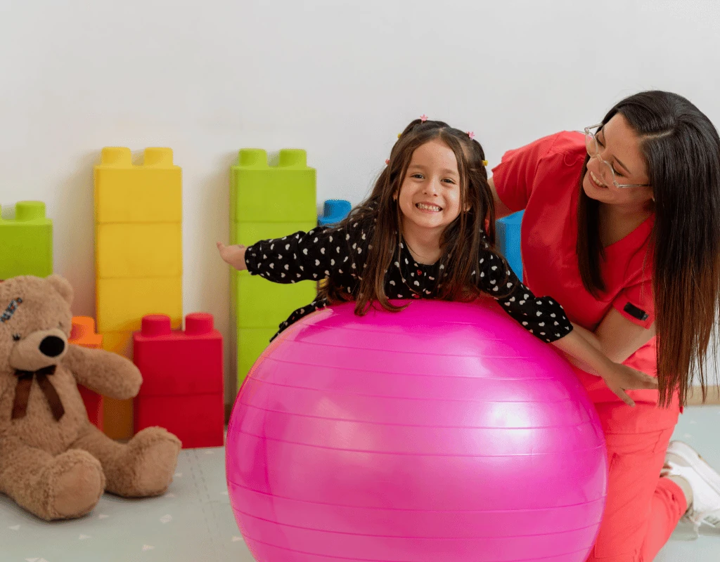 Therapist steadying a laughing young girl as she balances on a large pink therapy ball in a colourful playroom
