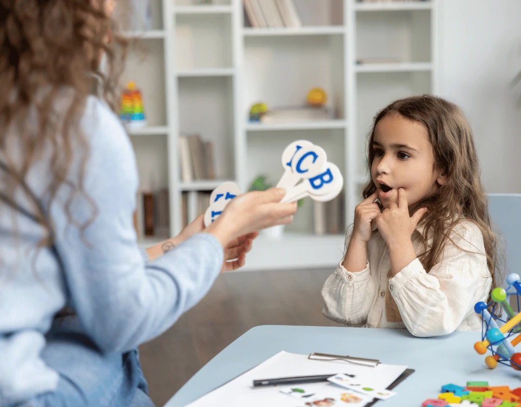 Speech therapist holding letter cards in front of a young girl seated at a table with learning materials