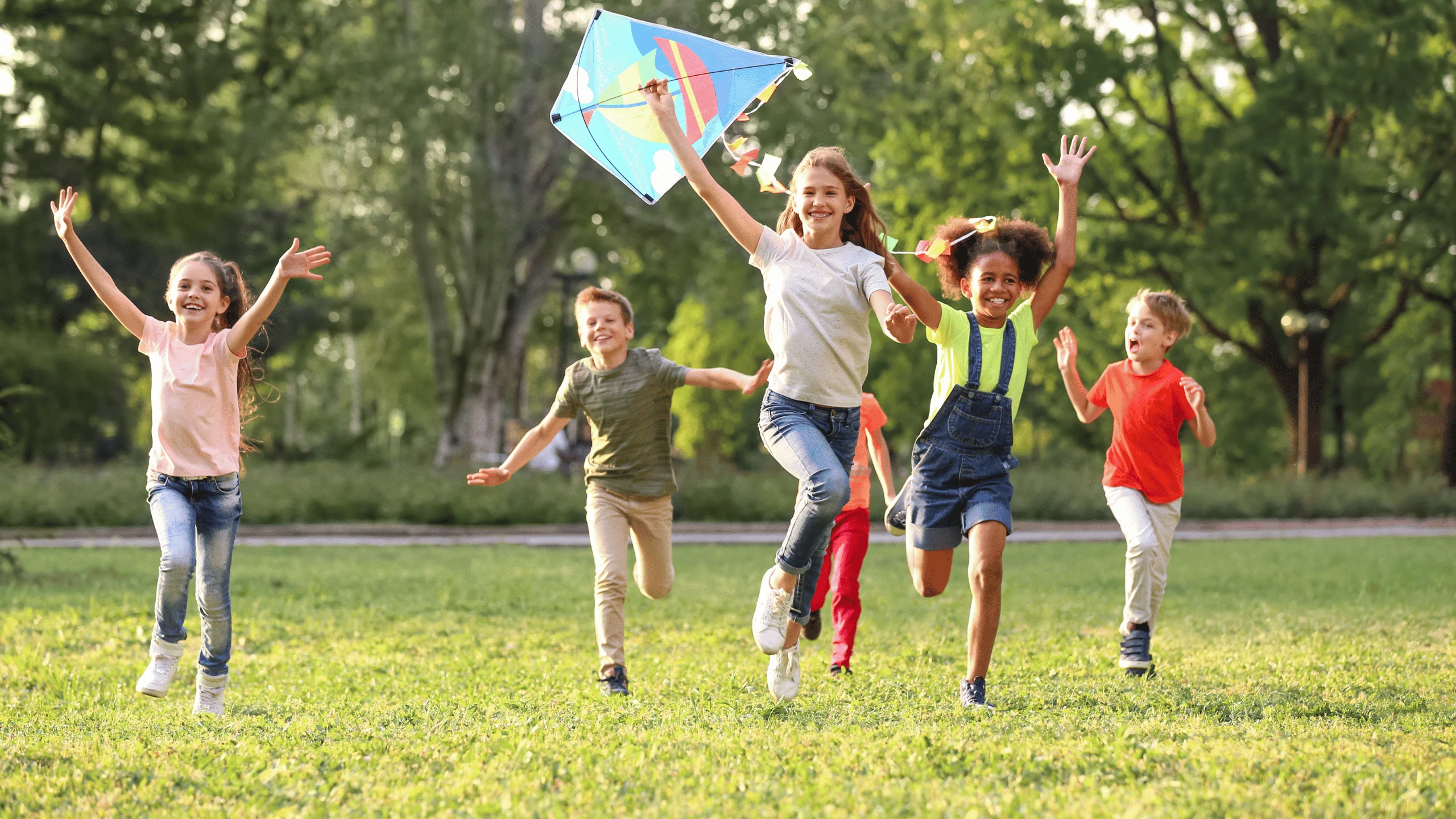 Four children running across grass together while flying a colourful kite