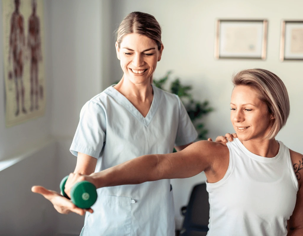 Therapist supporting a womans arm through a lateral dumbbell raise in a room with anatomy charts
