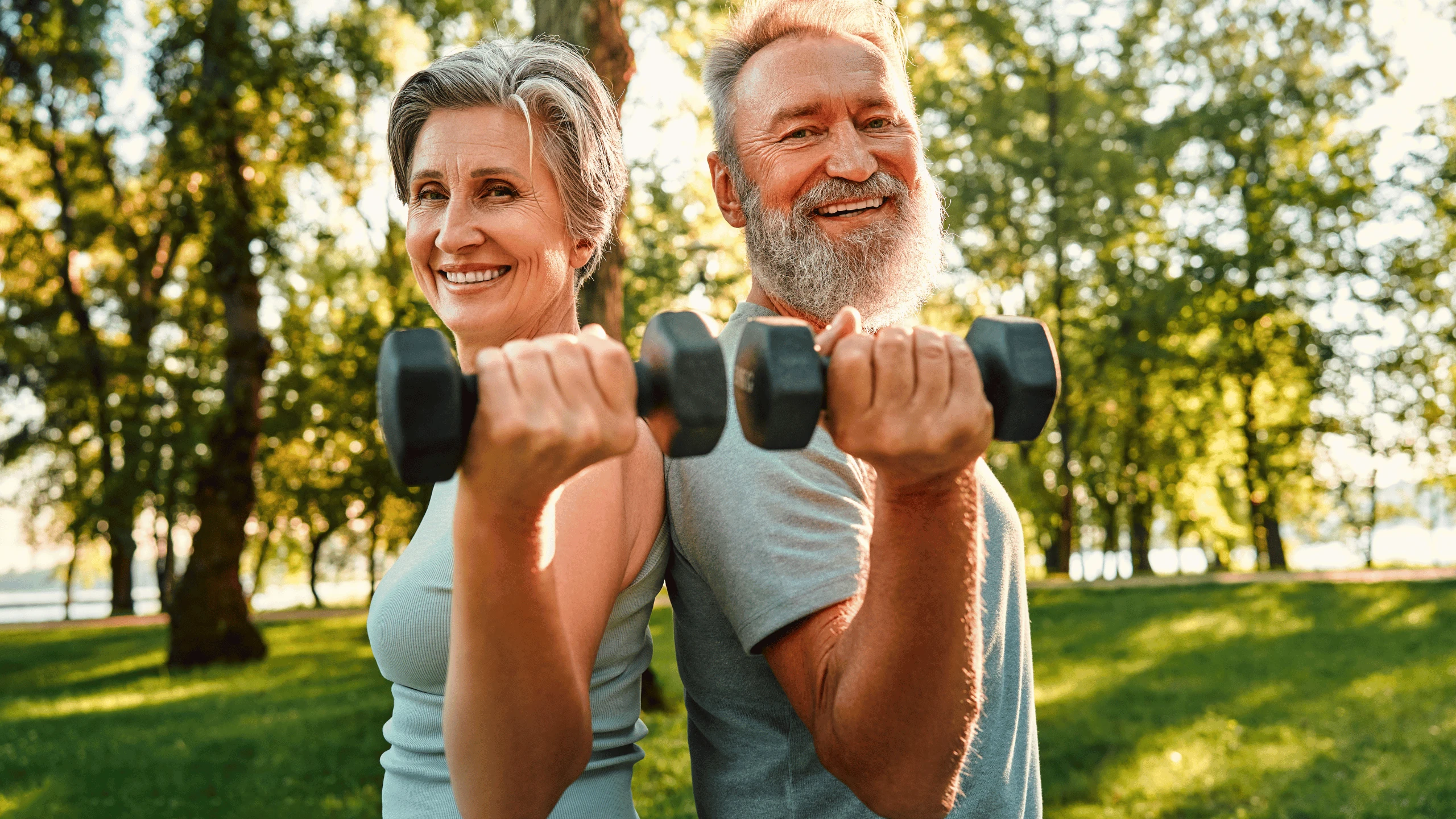 Older man and woman performing dumbbell curls together in a sunlit park