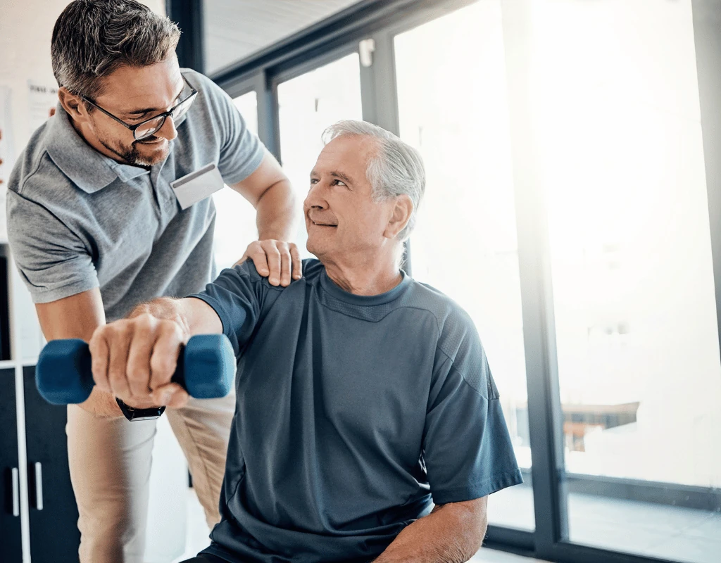 Therapist guiding an older man through a dumbbell shoulder exercise beside a bright window