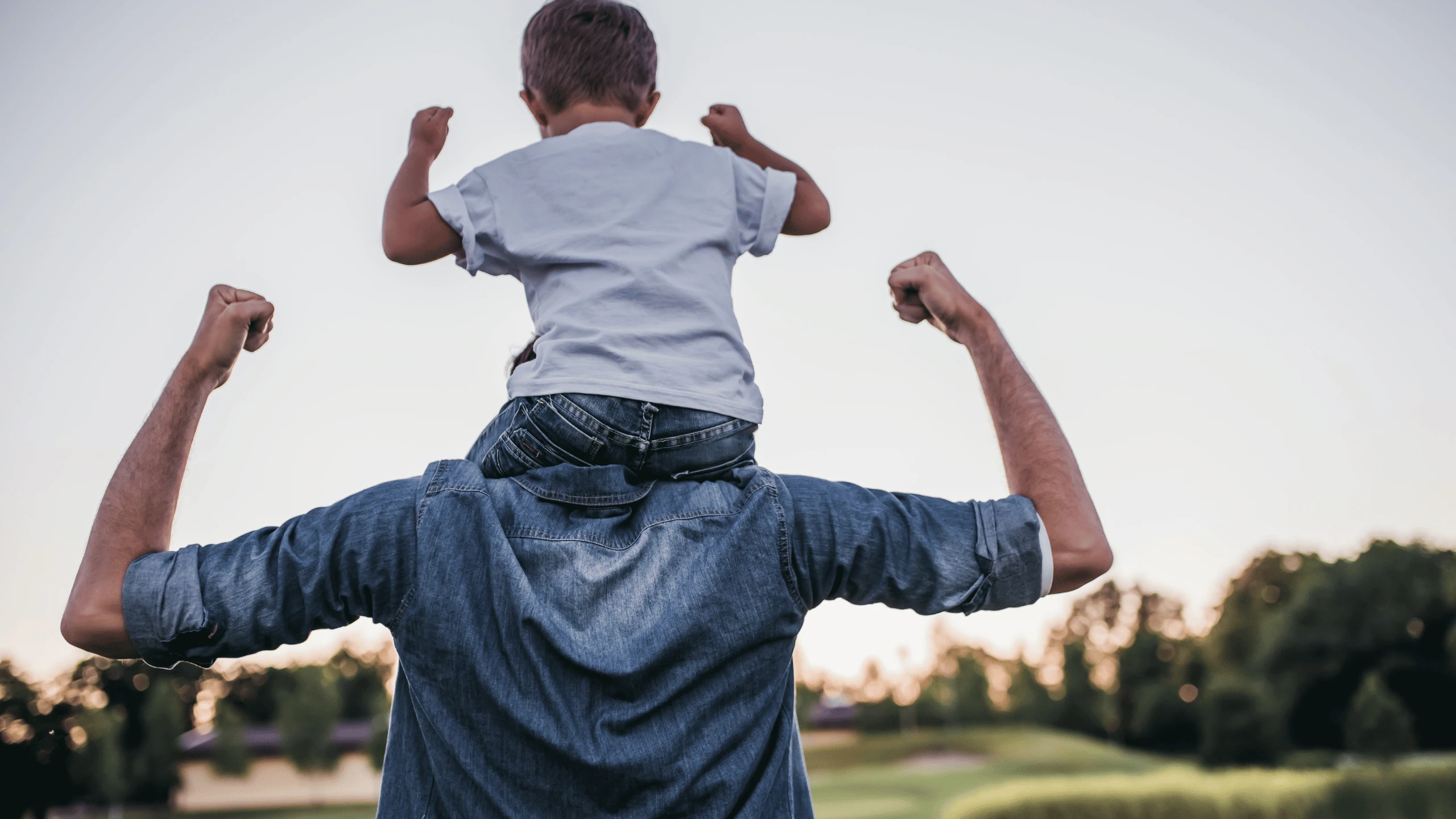 Man carrying a child on his shoulders outdoors, both flexing their arms, photographed from behind