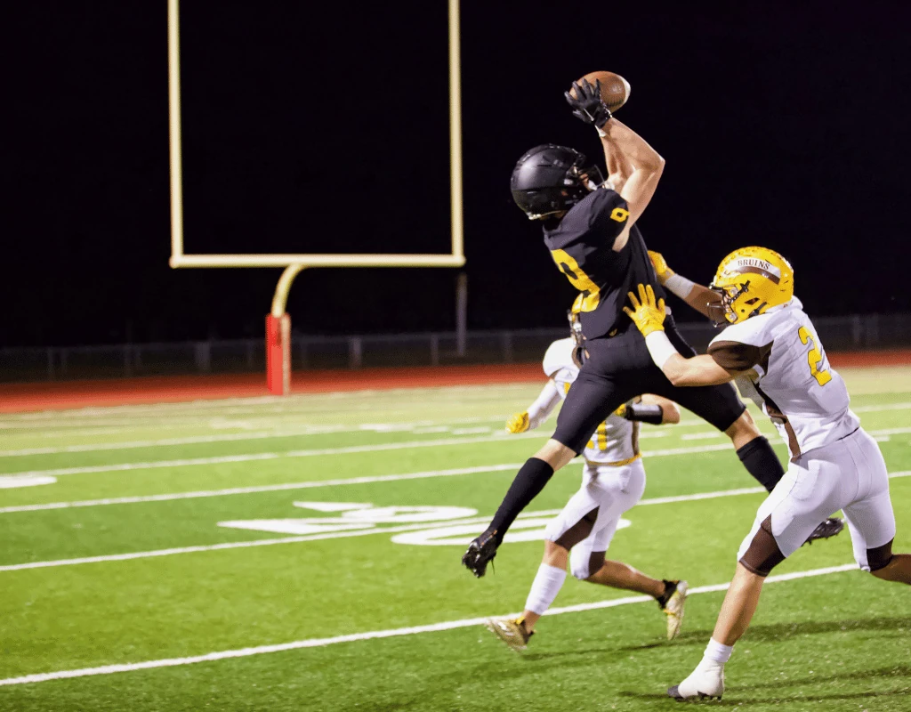 Football players in helmets and pads jumping for a ball near the goalposts under stadium lights