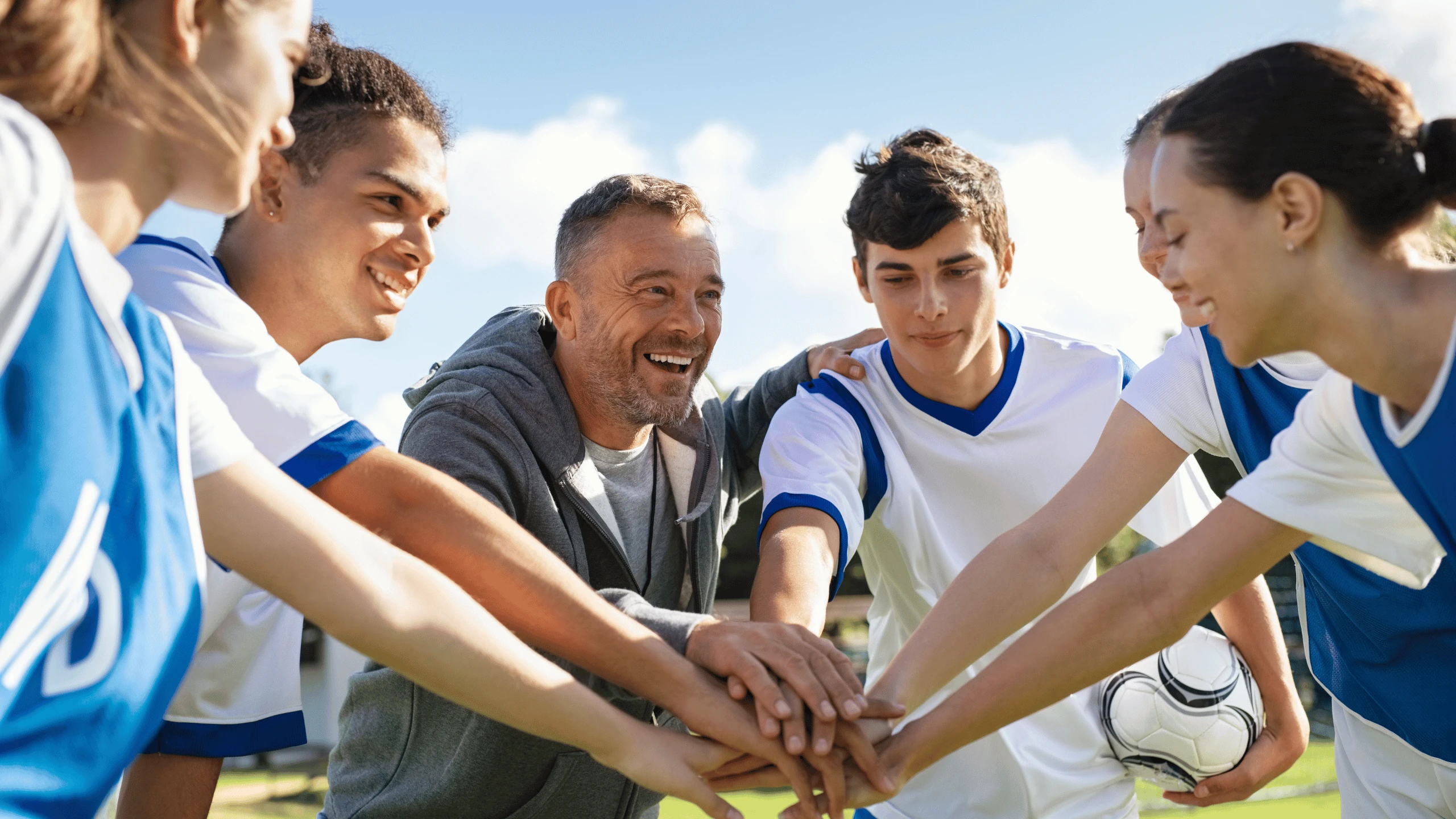 Young soccer players and a coach placing their hands together in a huddle beside a ball