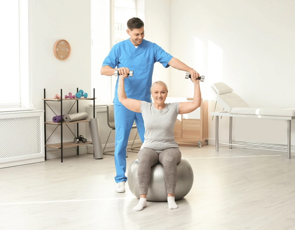 Therapist supporting a woman pressing weights overhead while she sits balanced on a grey exercise ball