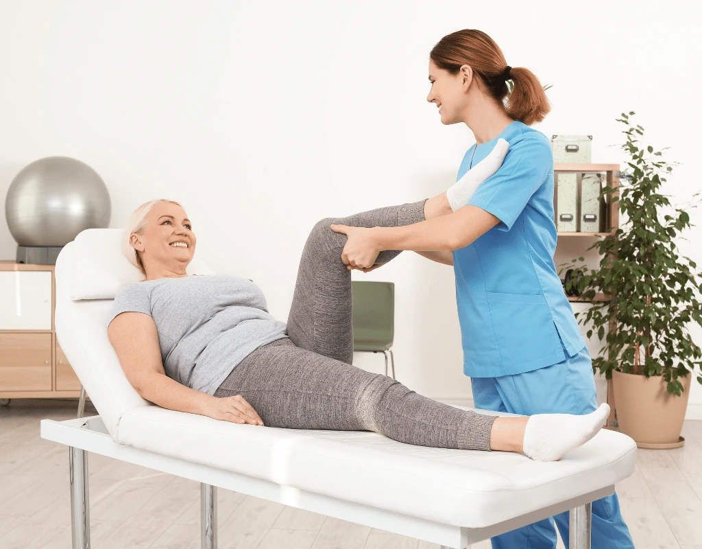 Therapist in blue scrubs moving an older womans leg while she lies on a treatment table