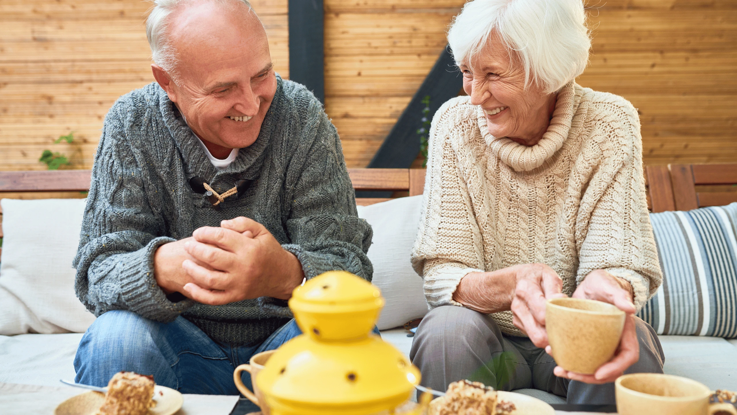 Older man and woman sitting together on a sofa with mugs and a yellow teapot on the table