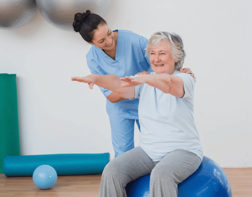 Therapist guiding an older womans arms forward while she sits balanced on a large therapy ball