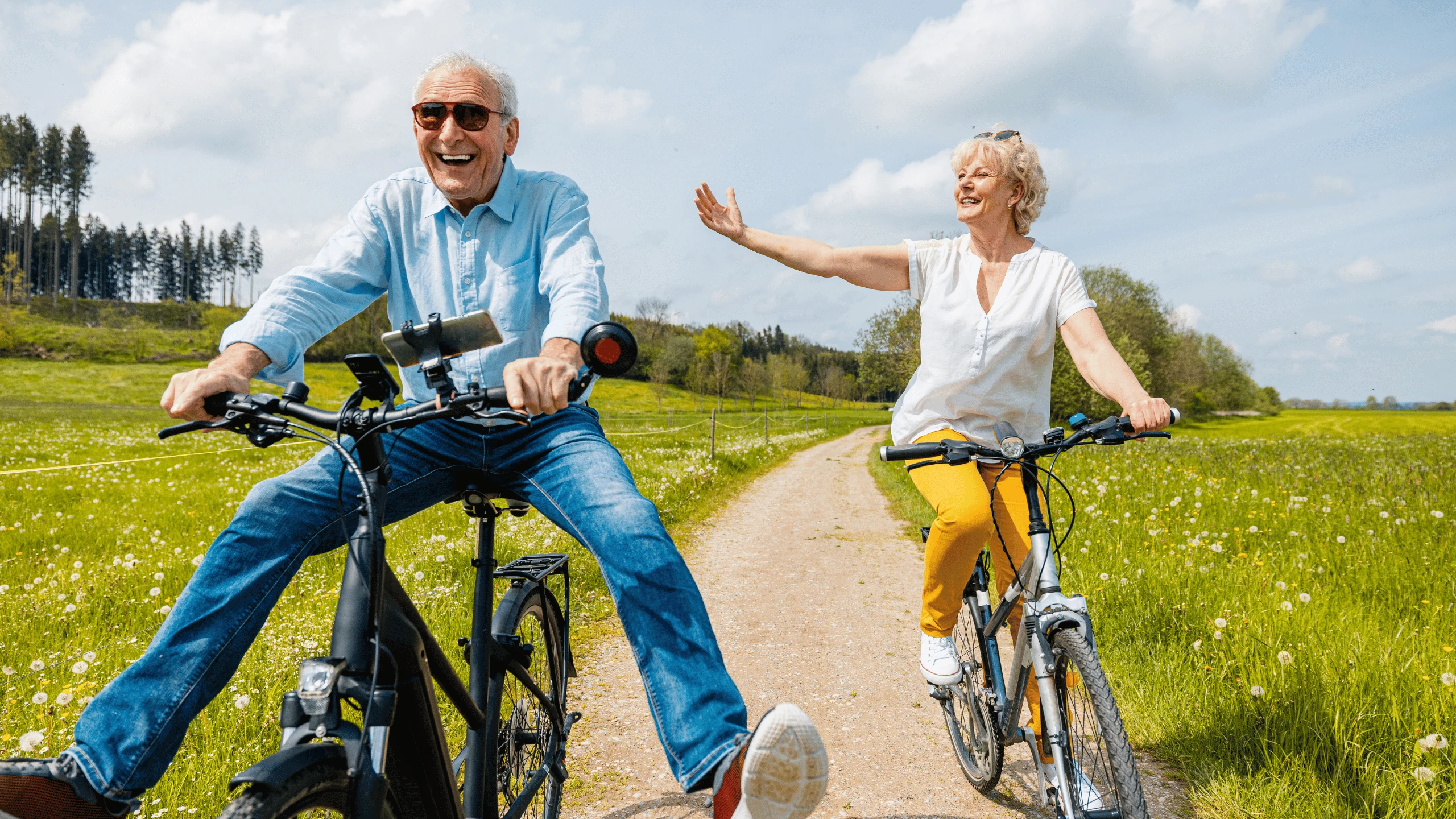 Older man and woman riding bicycles along a rural path, one raising a hand in greeting