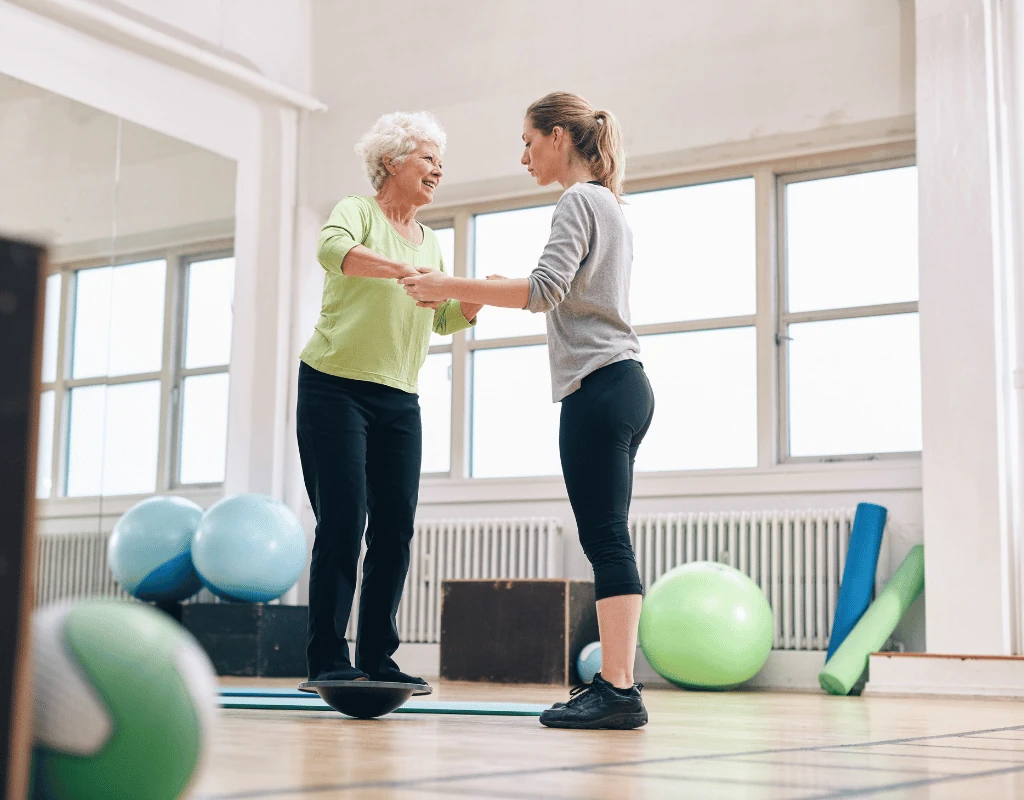 Therapist steadying an older woman as she balances on a wobble board in a bright therapy gym