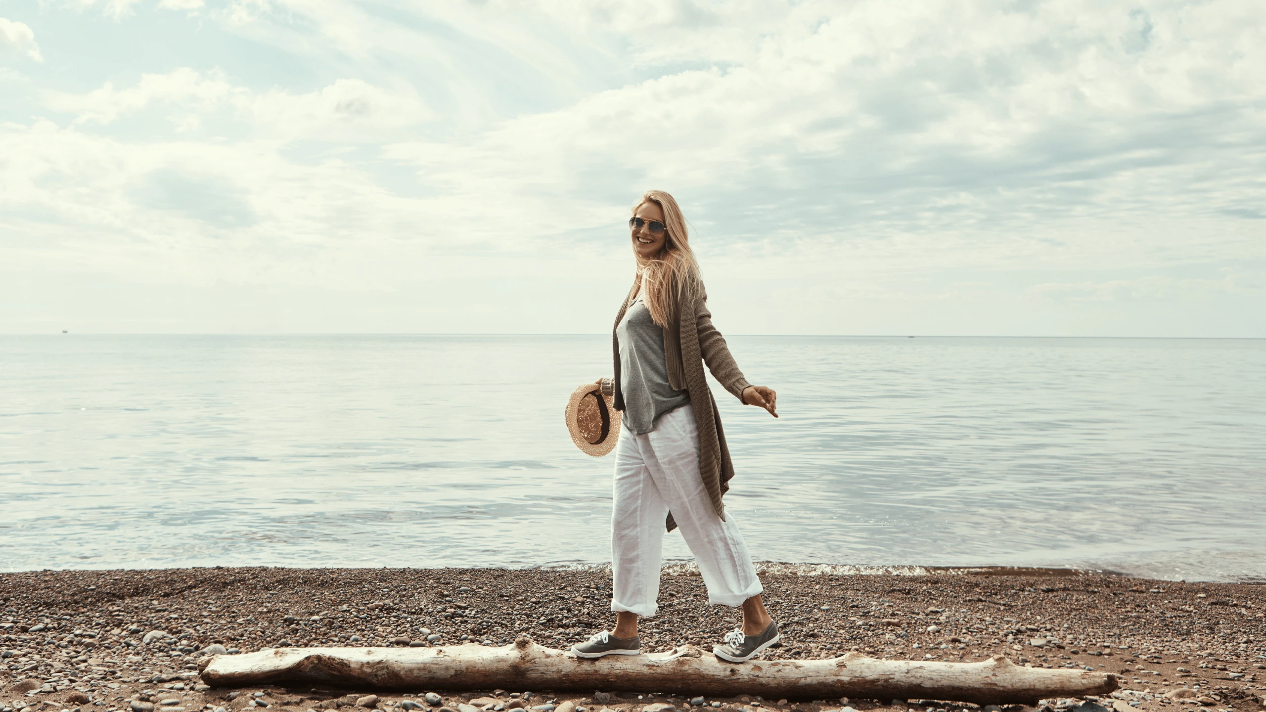 Woman walking along a beach at the edge of calm water under a pale sky