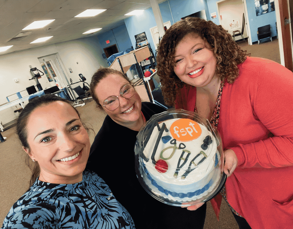 Three smiling colleagues holding a decorated cake carrying the clinic logo inside a therapy gym